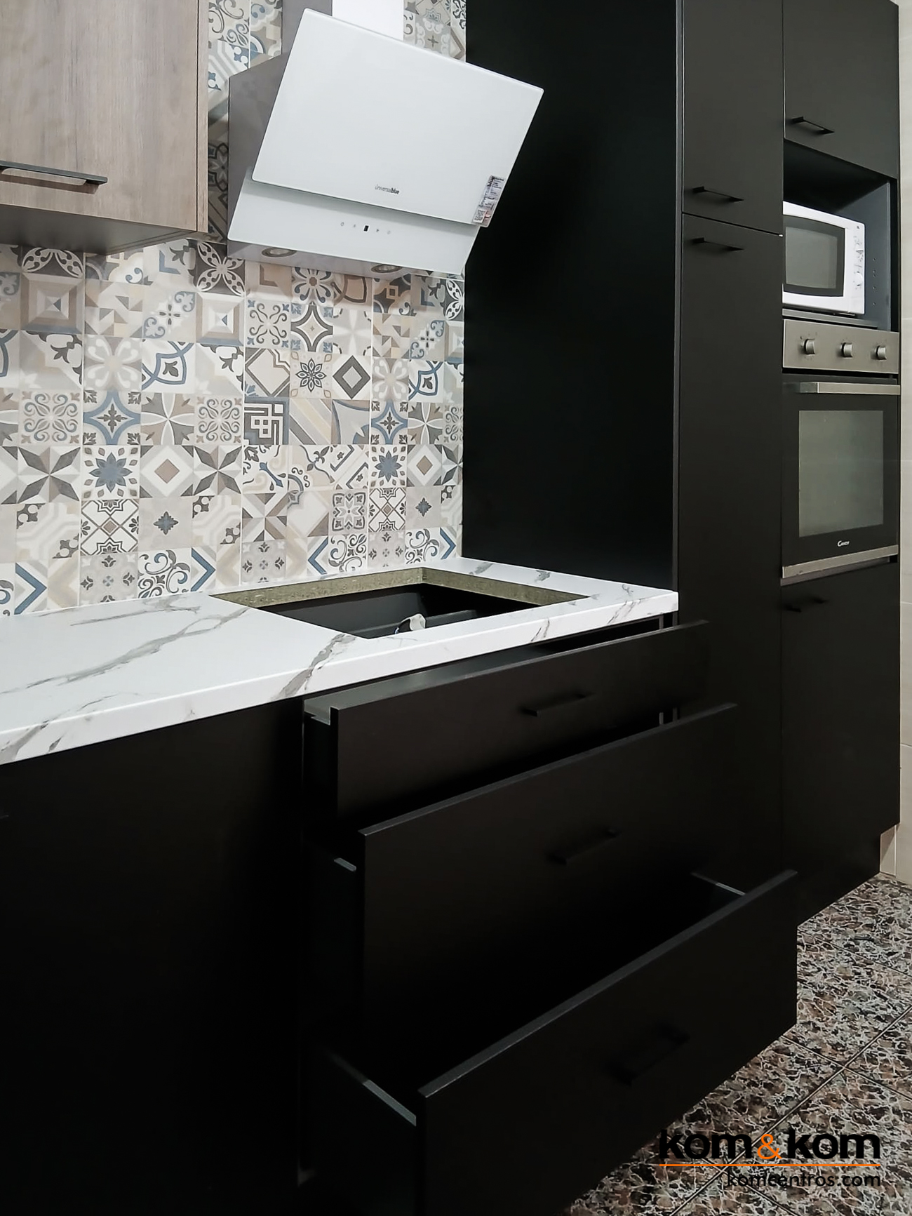 Cocina negra con encimera laminada marmolada, azulejos tipo hidráulico y campana inclinada blanca de pared.